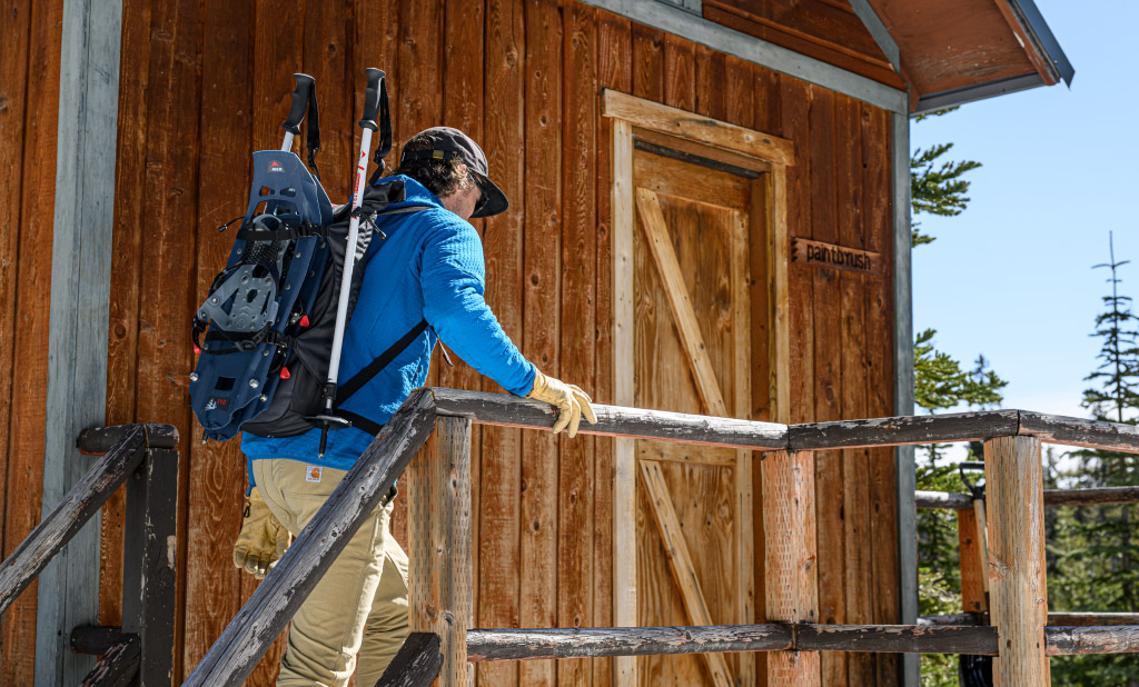 Man walking into cabin wearing Evo Trail Snowshoe Kit on his back